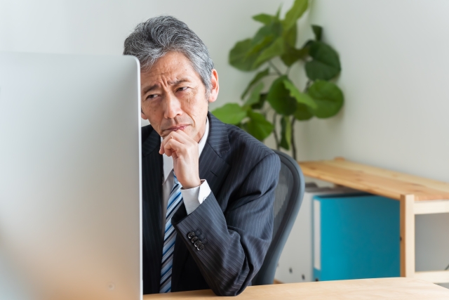 仕事をいつ、どこまで減らすか、職場の机で考え込む60歳前後の男性